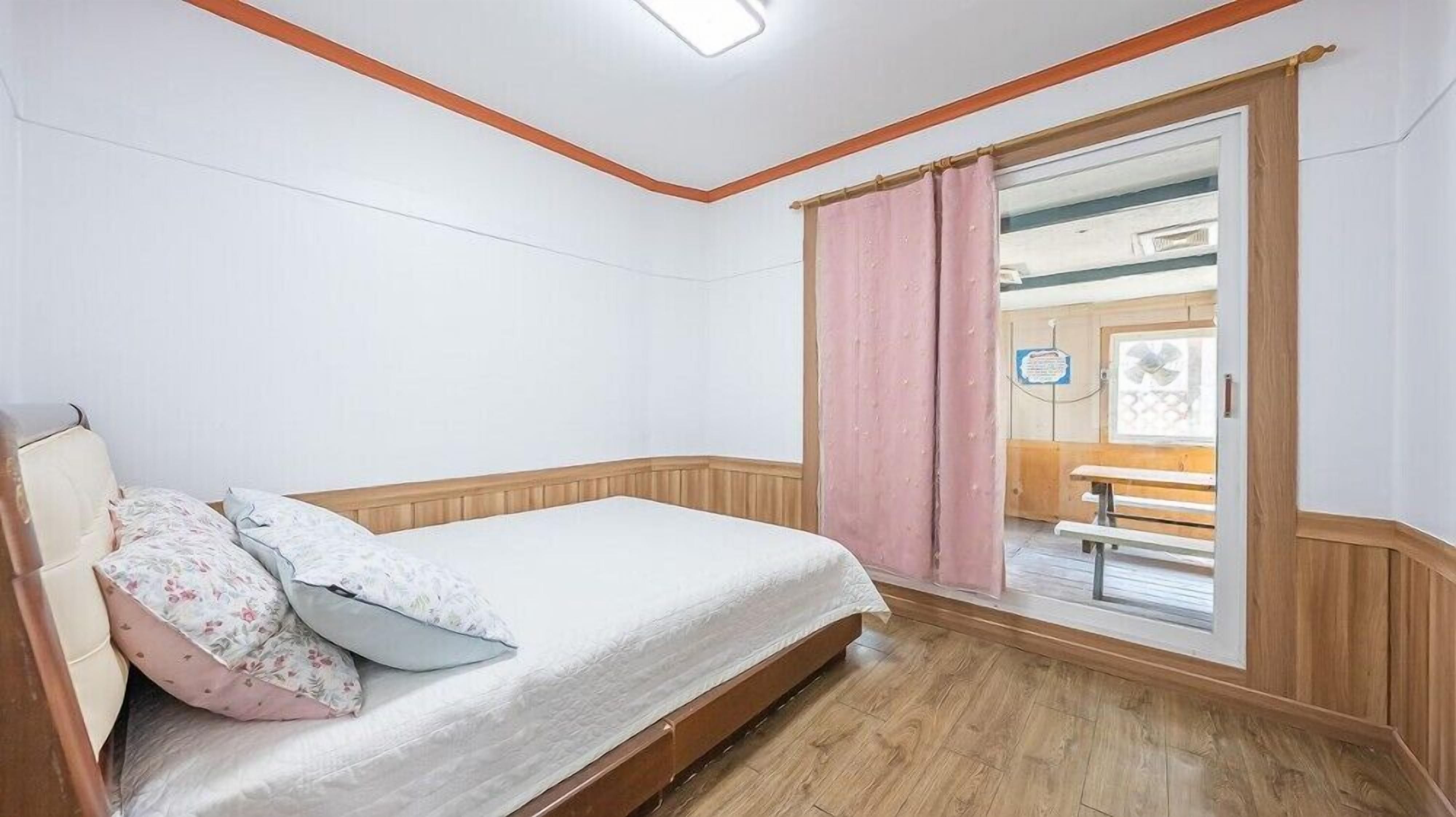 A single bed with a wooden headboard, a wooden floor, and a window with pink curtains.