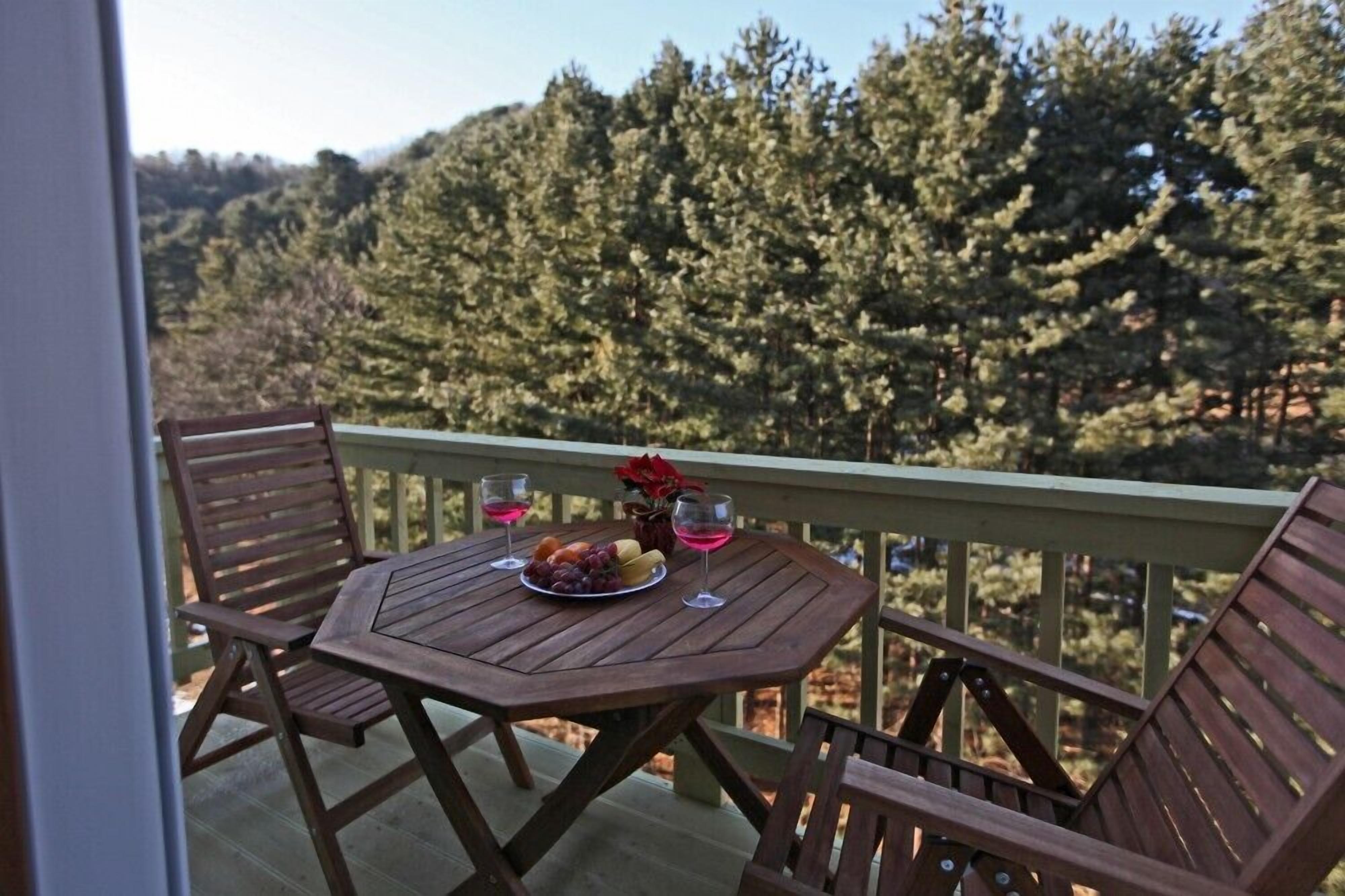 Una mesa de madera con un plato de fruta y dos vasos en un balcón con vista a un bosque.