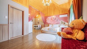 A bedroom with a large bed, a teddy bear, and a dining area in the background.