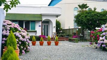 Un edificio con fachada blanca y contraventanas verdes, rodeado por un patio de grava con macetas y flores.