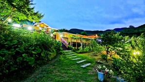 A well-lit outdoor area with a wooden staircase leading to a building, surrounded by lush greenery and a clear sky. - Sambatgol Pension (Pocheon)