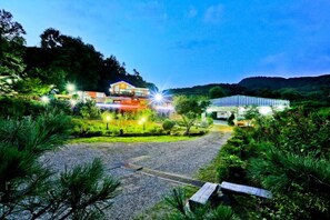 Un resort bien iluminado con entrada pavimentada, rodeado de exuberante vegetación y montañas al fondo.