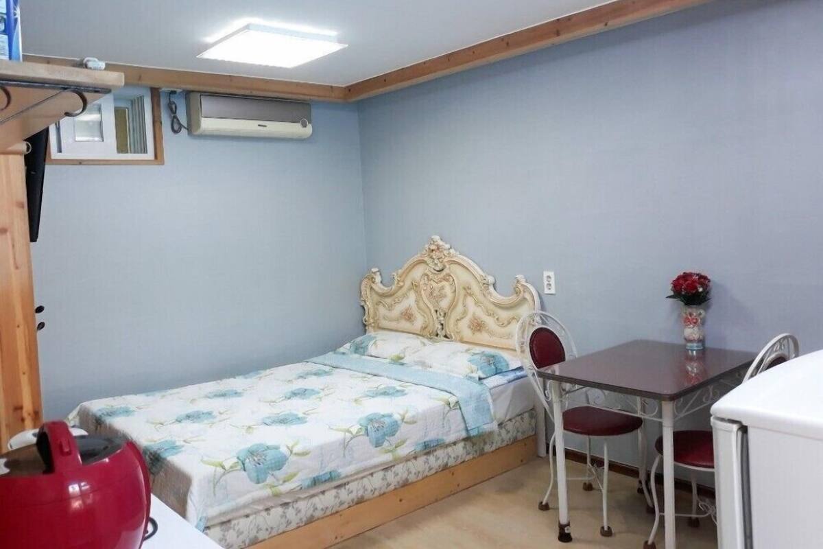 Quarto pequeno e compacto com uma cama de casal, uma mesa com cadeiras e uma área de cozinha compacta.
