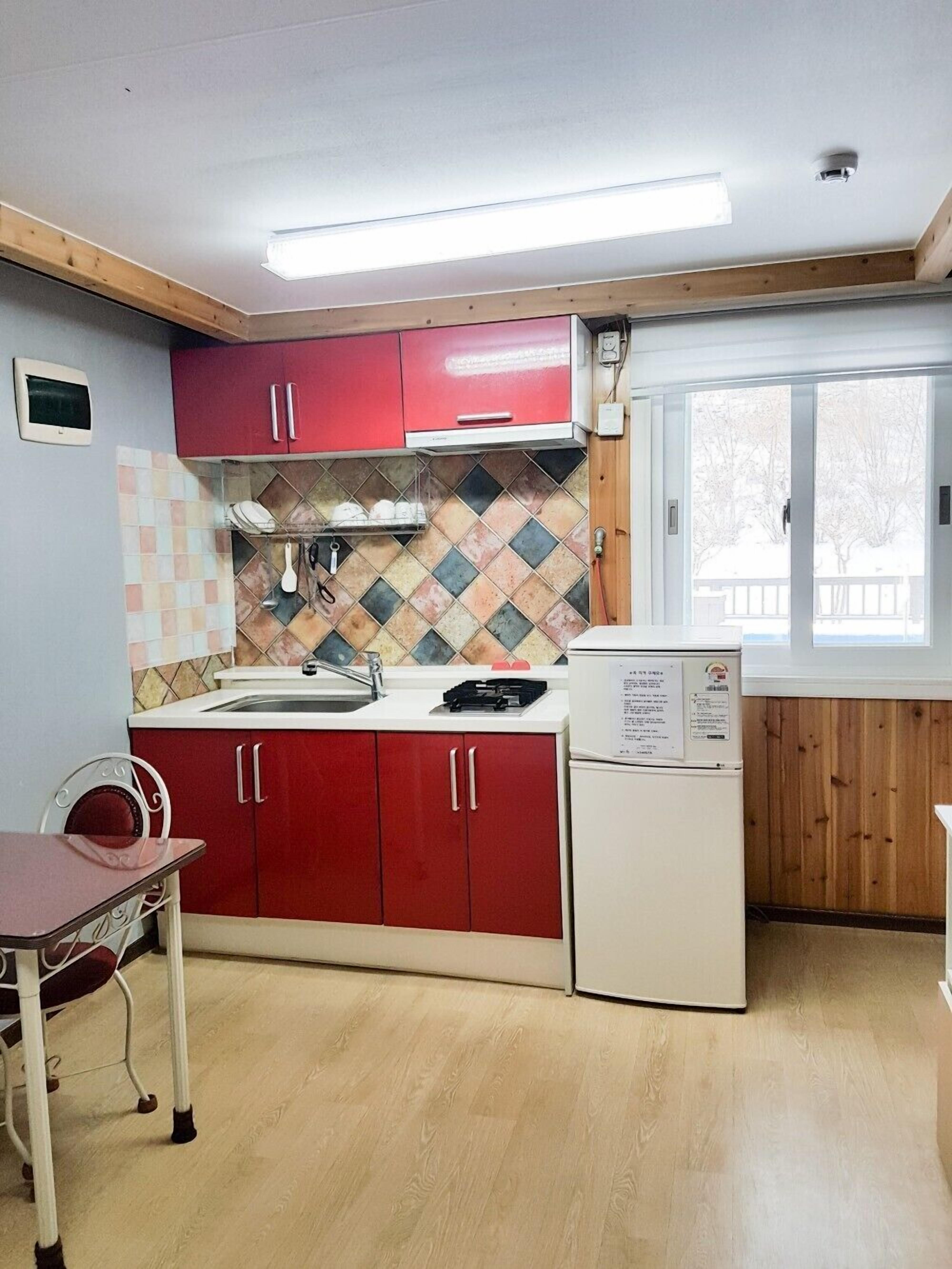 A compact kitchen with red cabinets, a tiled backsplash, a small sink, and a refrigerator.