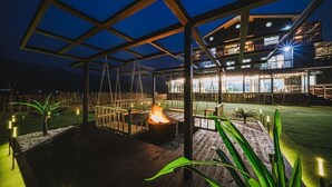 A modern outdoor patio with a fire feature, wooden deck, and a glass-enclosed building in the background. - Milky Way Pension (Gapyeong)