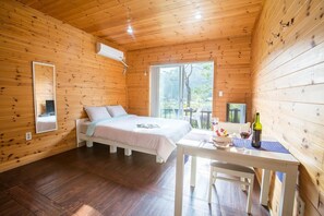 A wooden cabin room with a bed, a table with a bottle and glasses, and a window with a view of trees.