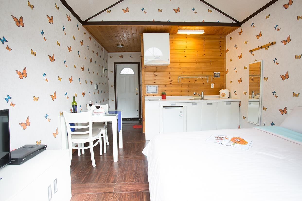 A bedroom with a bed, a desk with a chair, a small table with a bottle and a glass, and a kitchenette with a sink and cabinets.