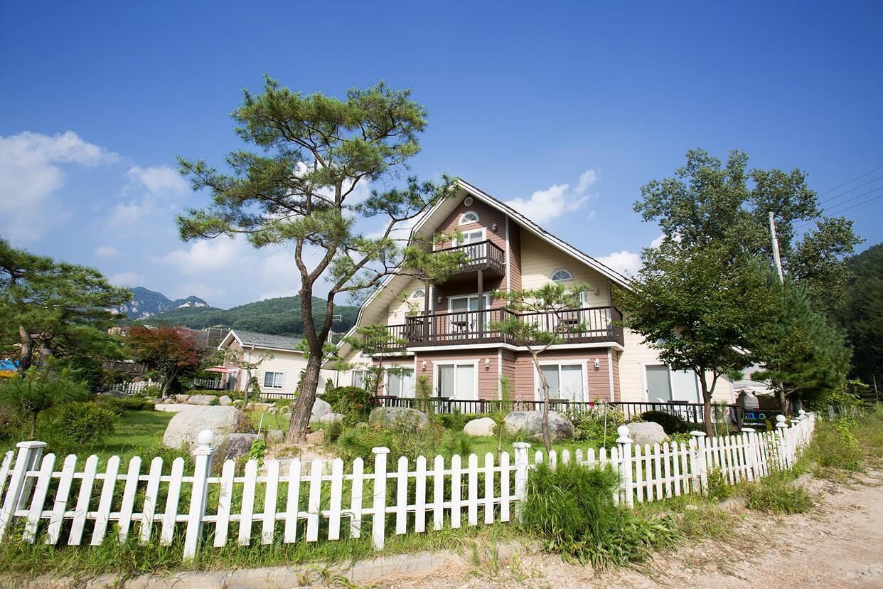 A two-story house with a balcony, surrounded by a white picket fence, trees, and a mountainous backdrop.
