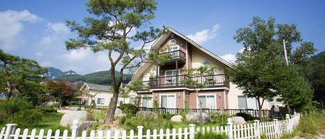 A two-story house with a balcony, surrounded by a white picket fence, trees, and a mountainous backdrop.
