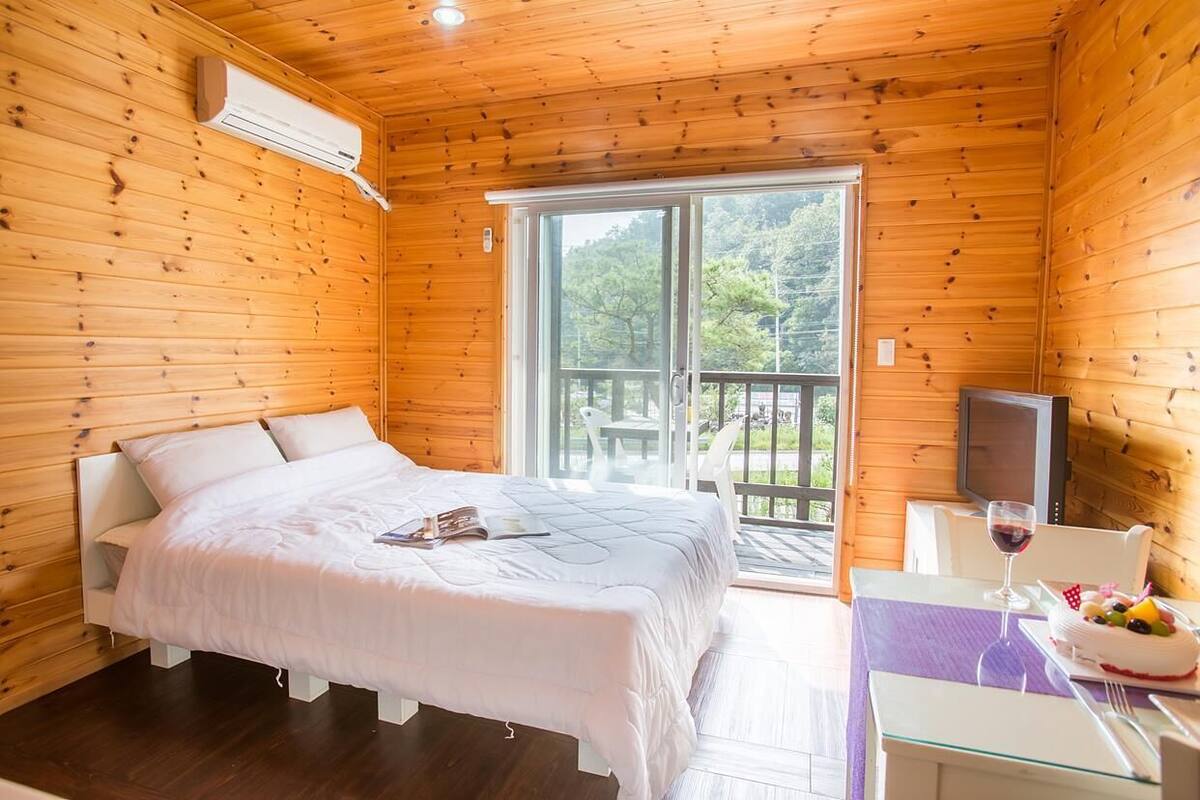 A wooden cabin room with a bed, a table with a glass top, and a TV.