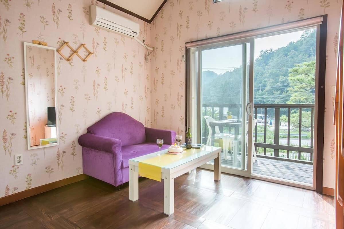 A room with a purple sofa, a glass table, and a large sliding glass door leading to a balcony with a view of greenery.