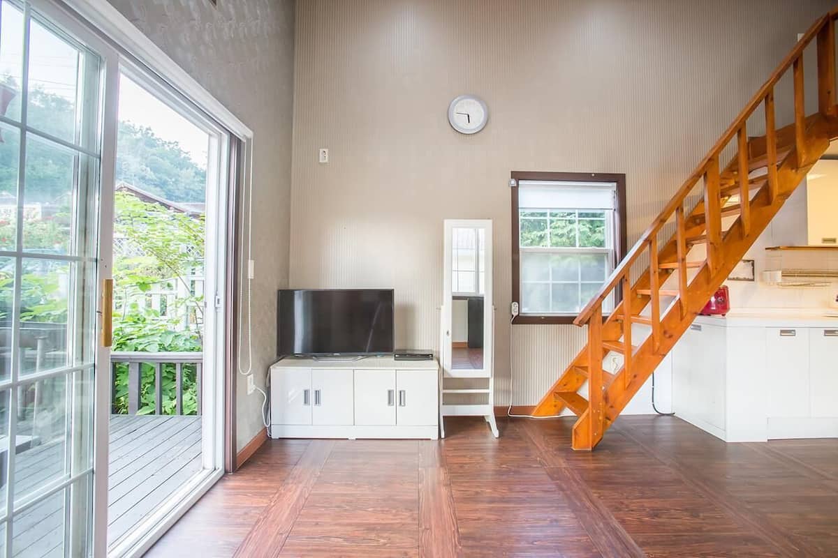 A spacious room with a wooden staircase, a television on a cabinet, and a large window offering a view of greenery.