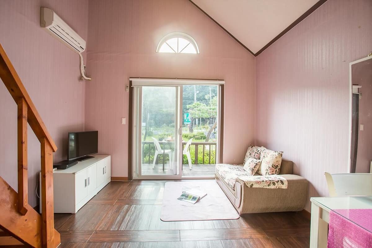 A room with a wooden floor, a sofa with floral cushions, a TV on a white cabinet, and a sliding glass door leading to an outdoor area.