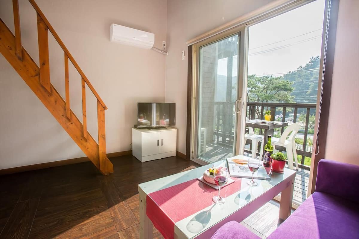 A room with a wooden staircase, a glass door leading to an outdoor area, a small white cabinet, and a table with a glass of wine and a bottle.