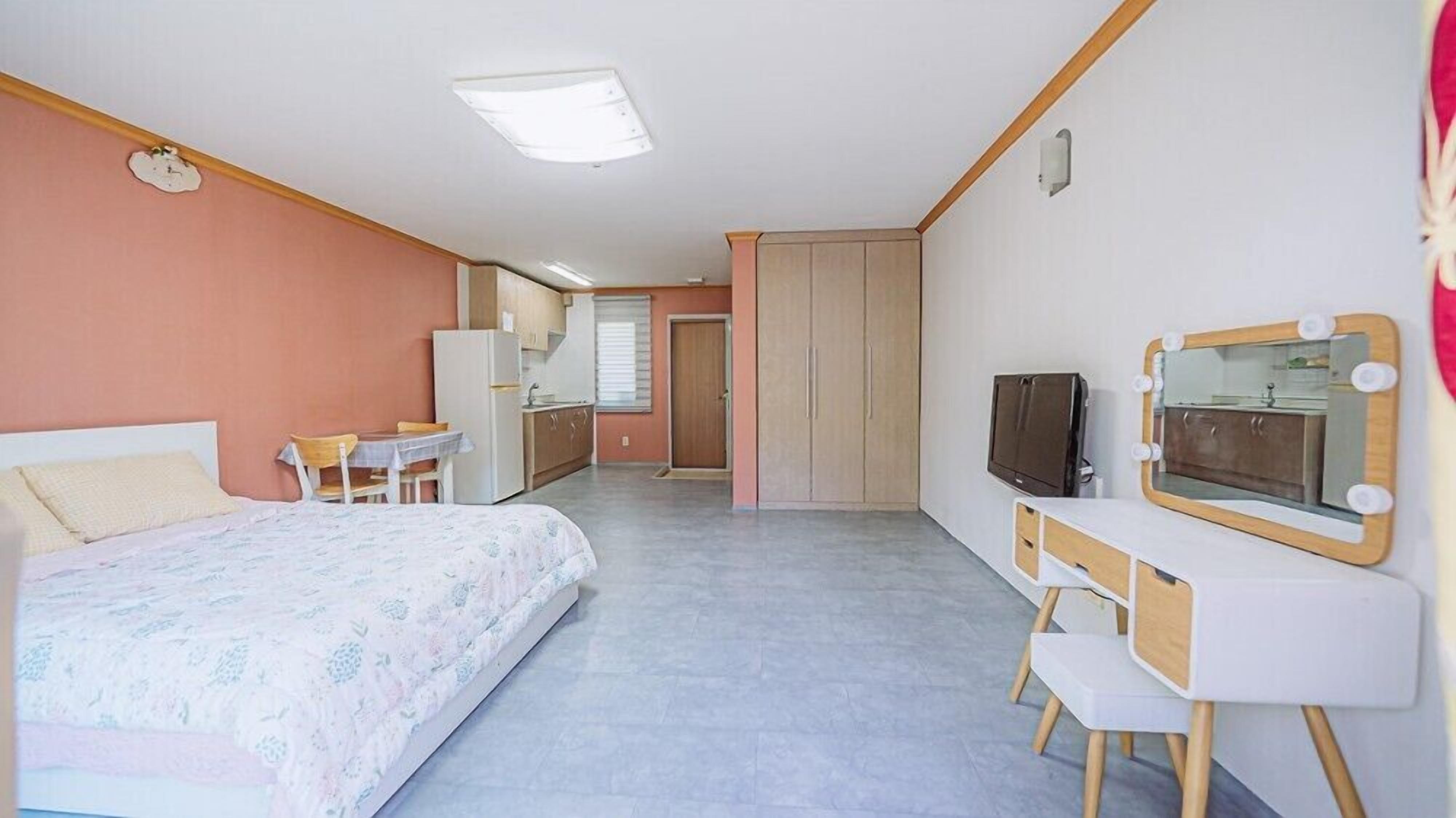 Ein kleines Hotelzimmer mit einem Bett, einer kleinen Küchenzeile, einem Schreibtisch mit Spiegel und einem Fernseher.