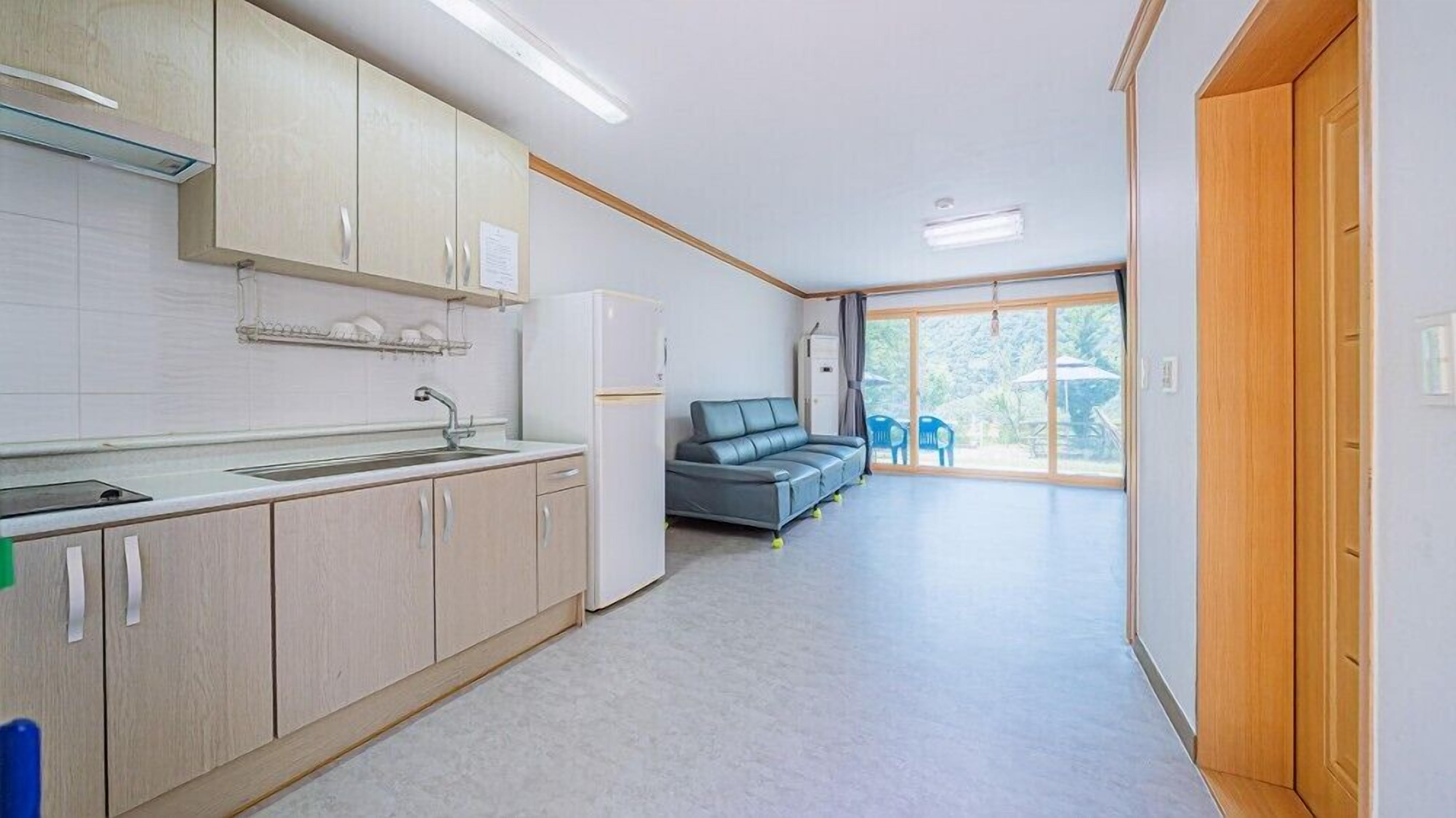 A modern kitchen with light wood cabinets, a built-in sink, and a refrigerator. There is a grey sofa and a dining area with blue chairs visible through a large window.