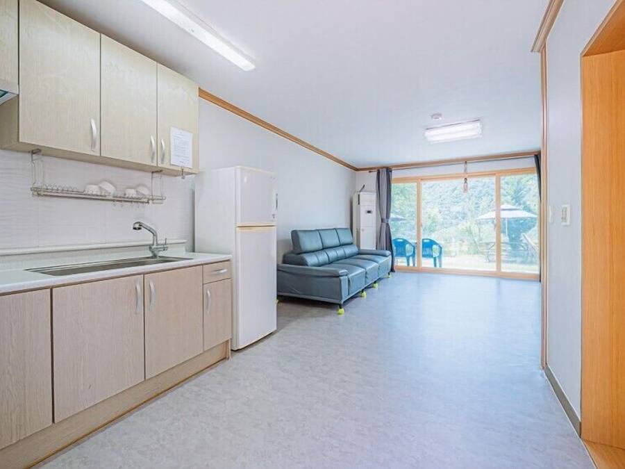 A modern kitchen with light wood cabinets, a built-in sink, and a refrigerator. There is a grey sofa and a dining area with blue chairs visible through a large window.