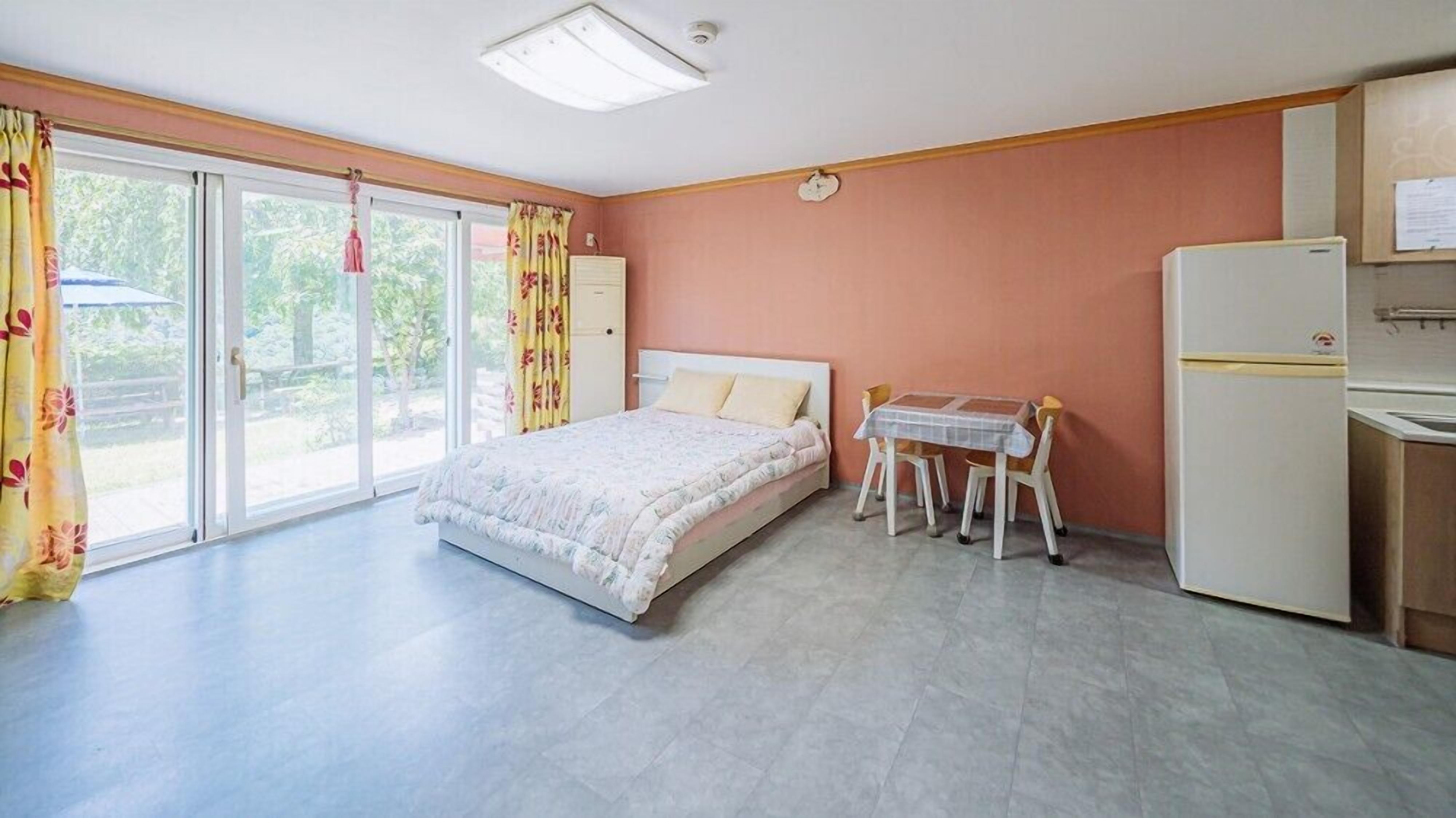 Un dormitorio con una cama, una mesita con sillas, un refrigerador y una ventana con cortinas.