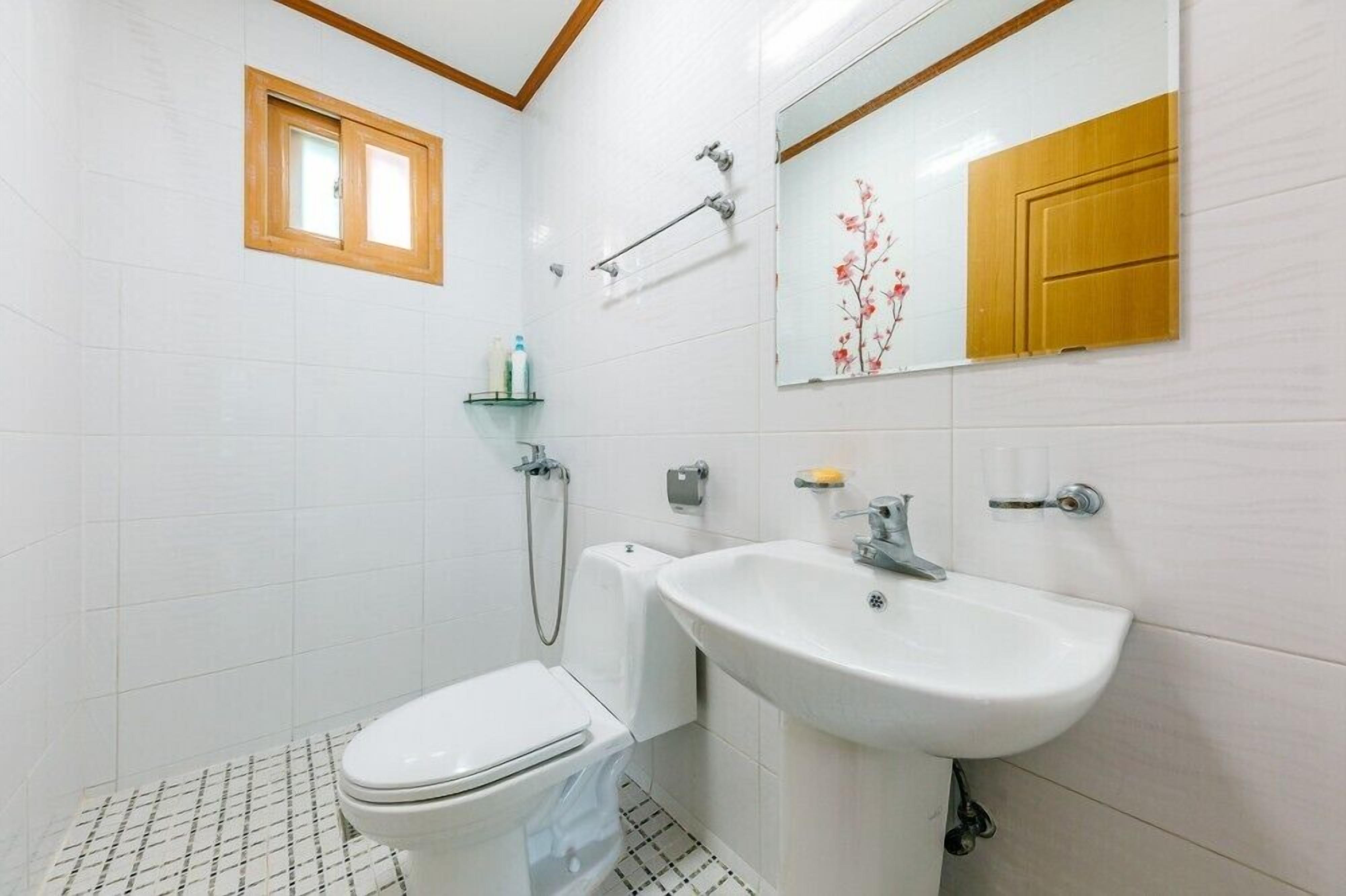 Une petite salle de bain propre, équipée de toilettes, d’un lavabo et d’une douche. Il y a une fenêtre au-dessus des toilettes et un miroir au-dessus du lavabo.