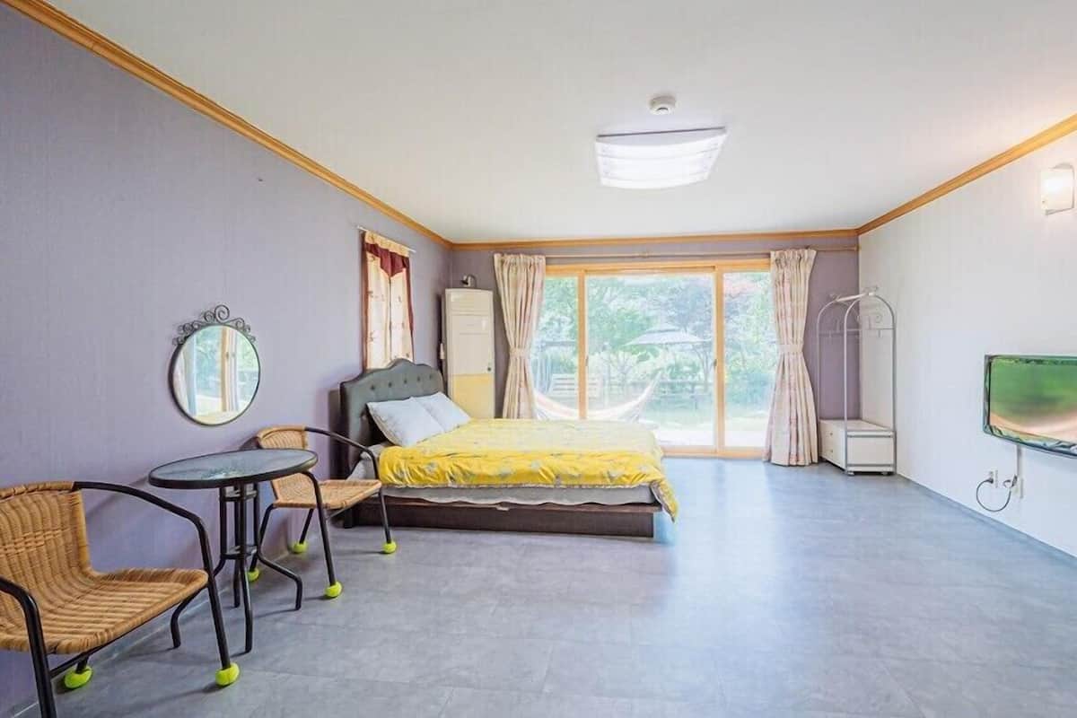 A bedroom with a bed, a chair, a small table, a mirror, a TV, and a window with curtains.
