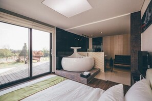 A modern bedroom with a large bed, a built-in bathtub, and a view of a deck and trees. - Terate Pension (Chuncheon)
