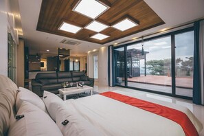 Un dormitorio moderno con una cama grande, un sofá y una mesa de comedor. Hay un ventanal corredero que da a un balcón con vistas al exterior.