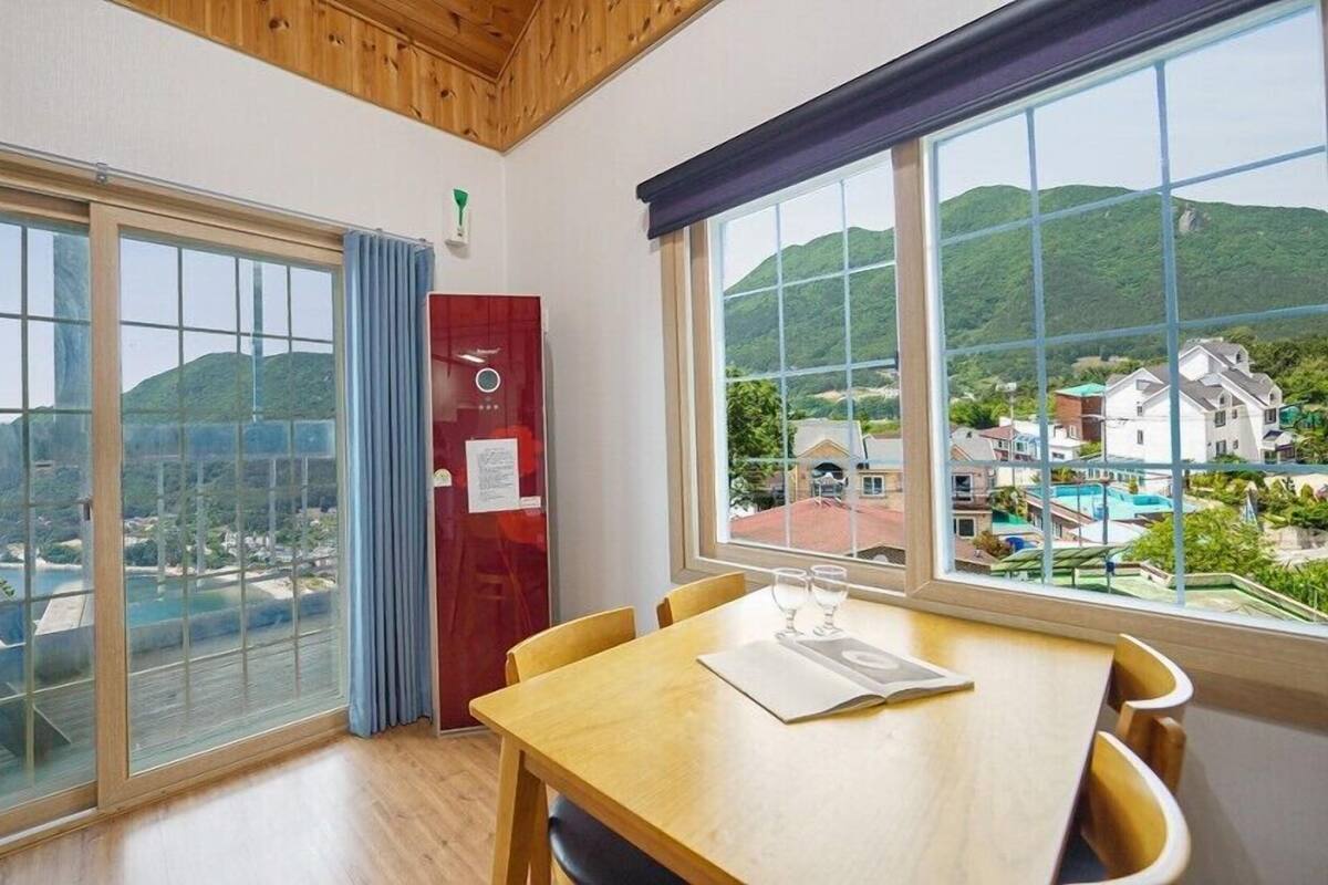 A room with large windows offering a view of a beach and hills, a wooden table with a magazine, and a red cabinet.