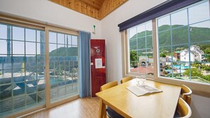 A room with large windows offering a view of a beach and hills, a wooden table with a magazine, and a red cabinet. - Geoje Cheongsol Pension (Geoje)