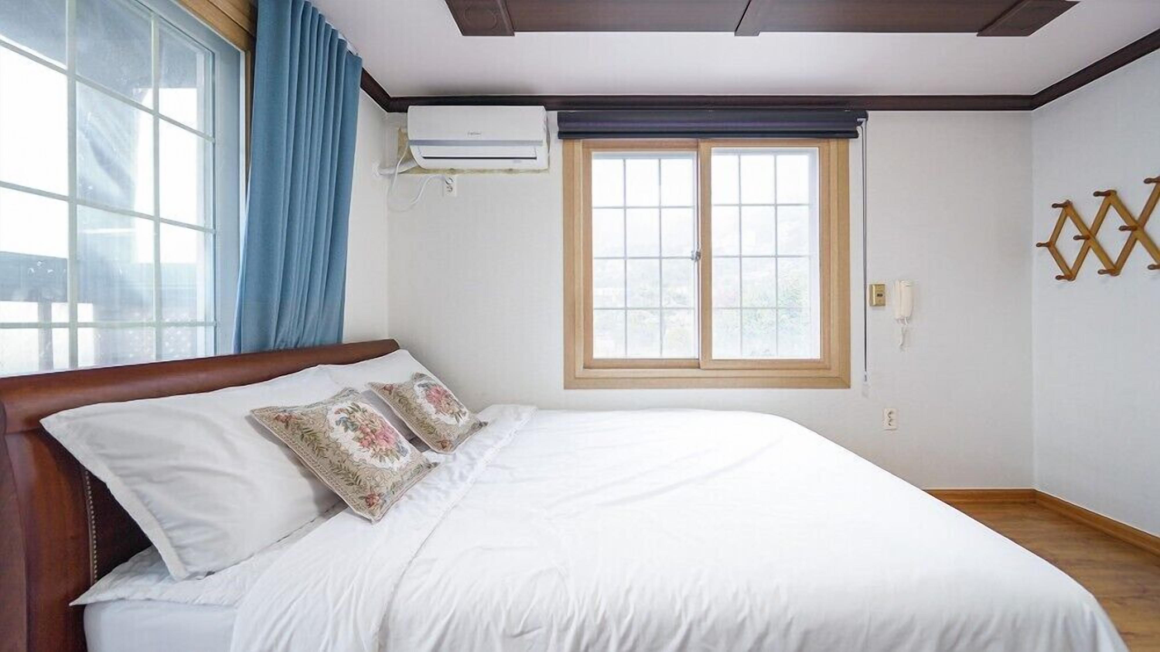 A bedroom with a wooden bed, white bedding, a window with a wooden frame, and an air conditioning unit on the wall.