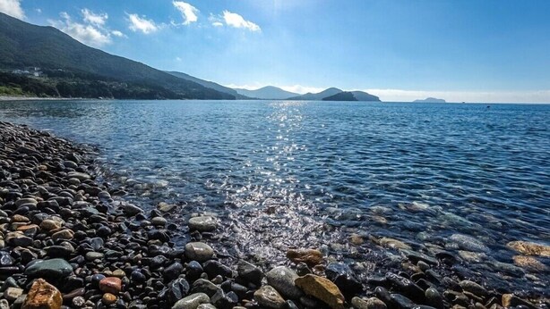 A clear lake with a rocky shoreline and distant mountains under a bright sun. - Geoje Cheongsol Pension (Geoje)