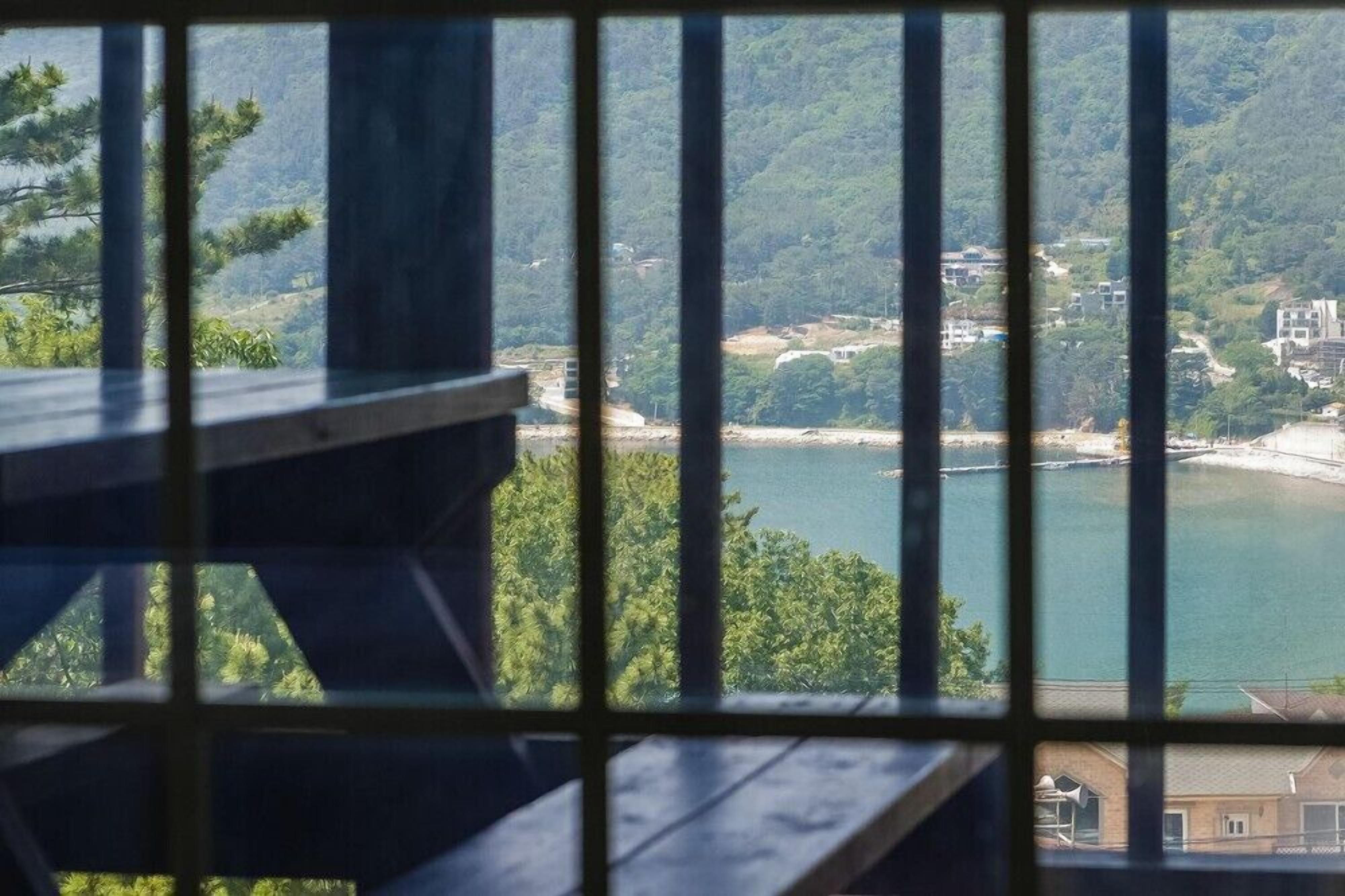 A view through a window with a metal grid, overlooking a body of water, a forested hillside, and some buildings in the distance.