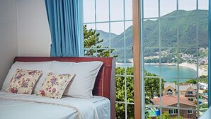 A bedroom with a bed, floral pillows, and a large window offering a view of a coastal landscape. - Geoje Cheongsol Pension (Geoje)