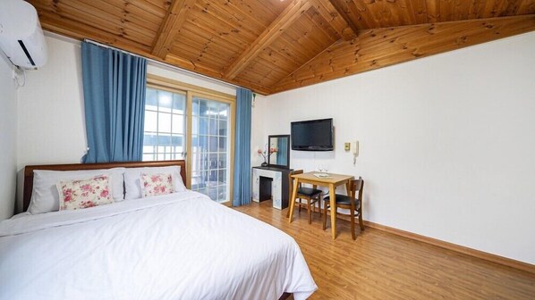 A bedroom with a wooden ceiling, a bed with floral pillows, a desk with a chair, and a TV on the wall. - Geoje Cheongsol Pension (Geoje)