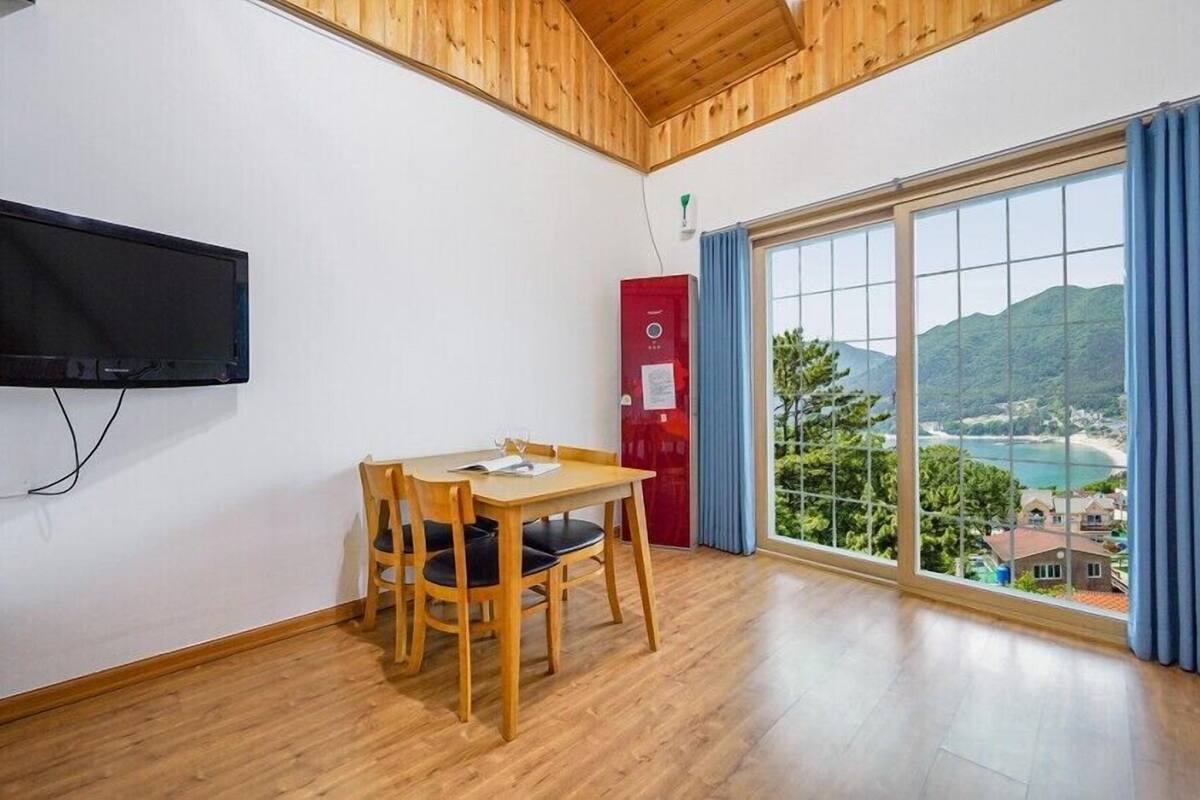 A room with a wooden ceiling, a TV mounted on the wall, a dining table with chairs, a red cabinet, and a large window offering a scenic view.