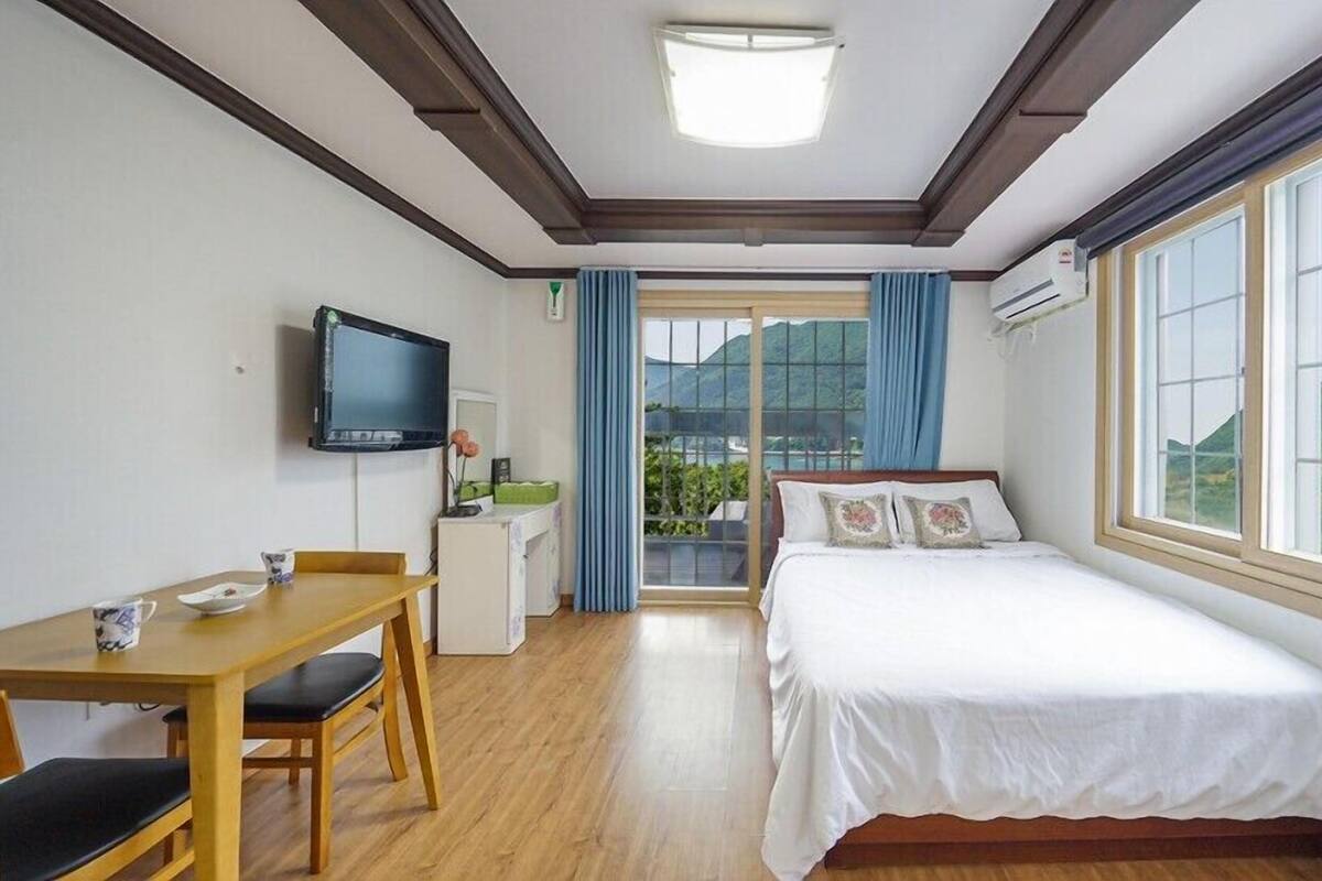 A hotel room with a bed, a desk, a TV, and a window with a view of greenery.