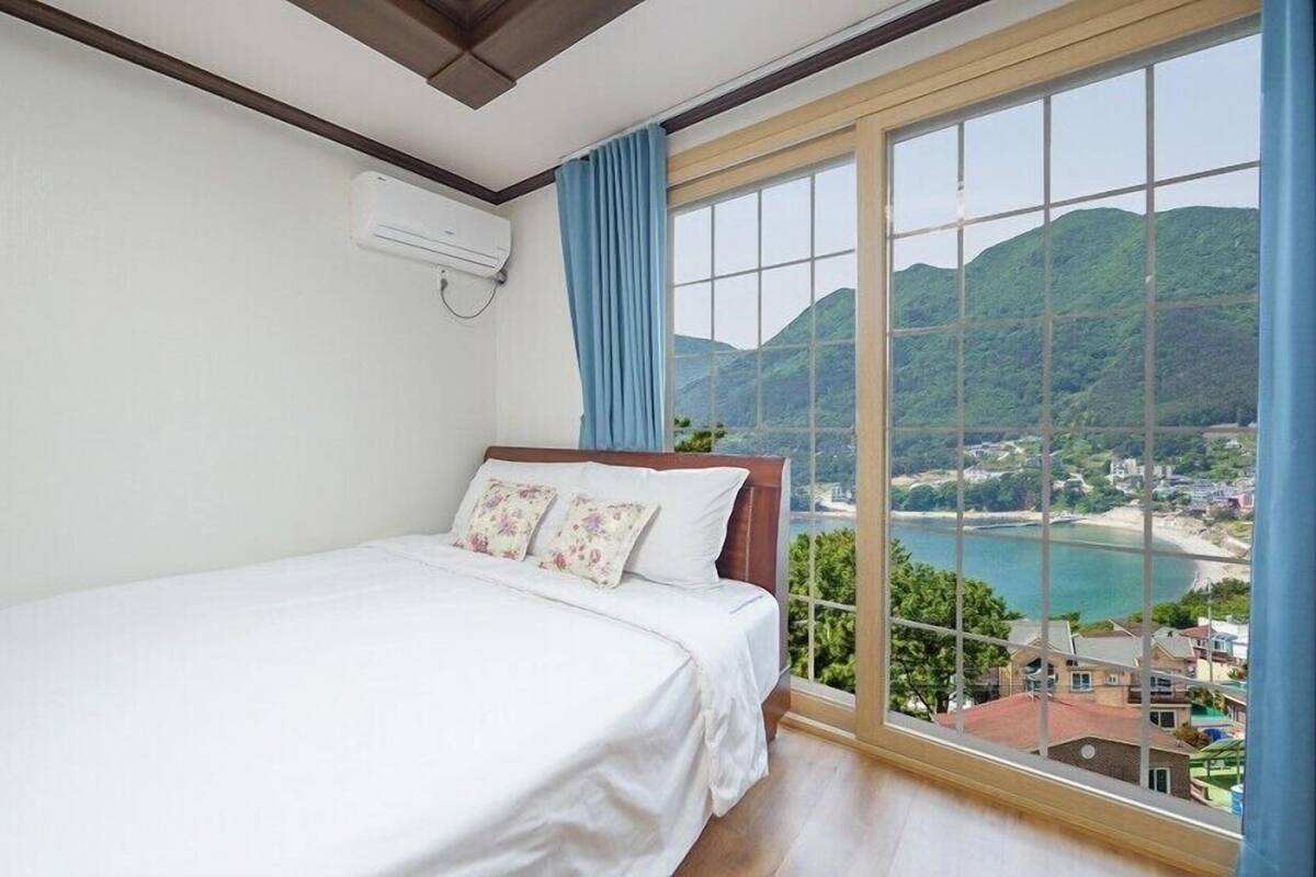 A bedroom with a bed, a window with a view of a coastal area, and an air conditioning unit on the wall.