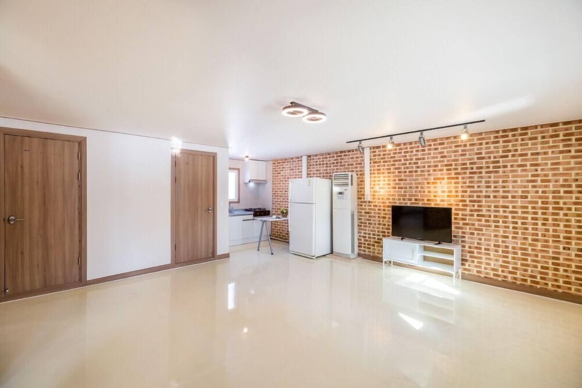 A spacious living area with a kitchen, a TV on a stand, and a brick wall.