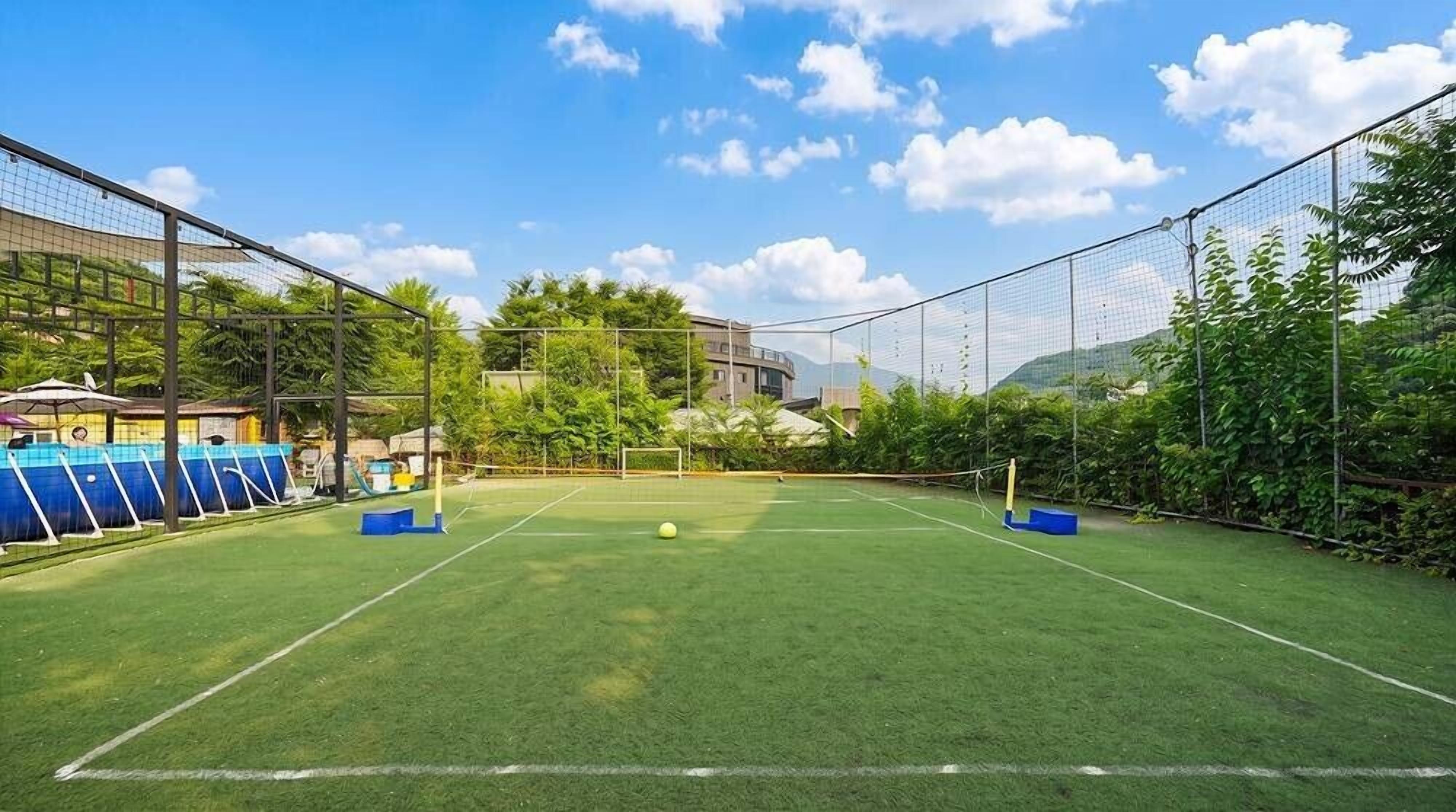 An outdoor tennis court with a net, surrounded by a fence, and a clear sky.