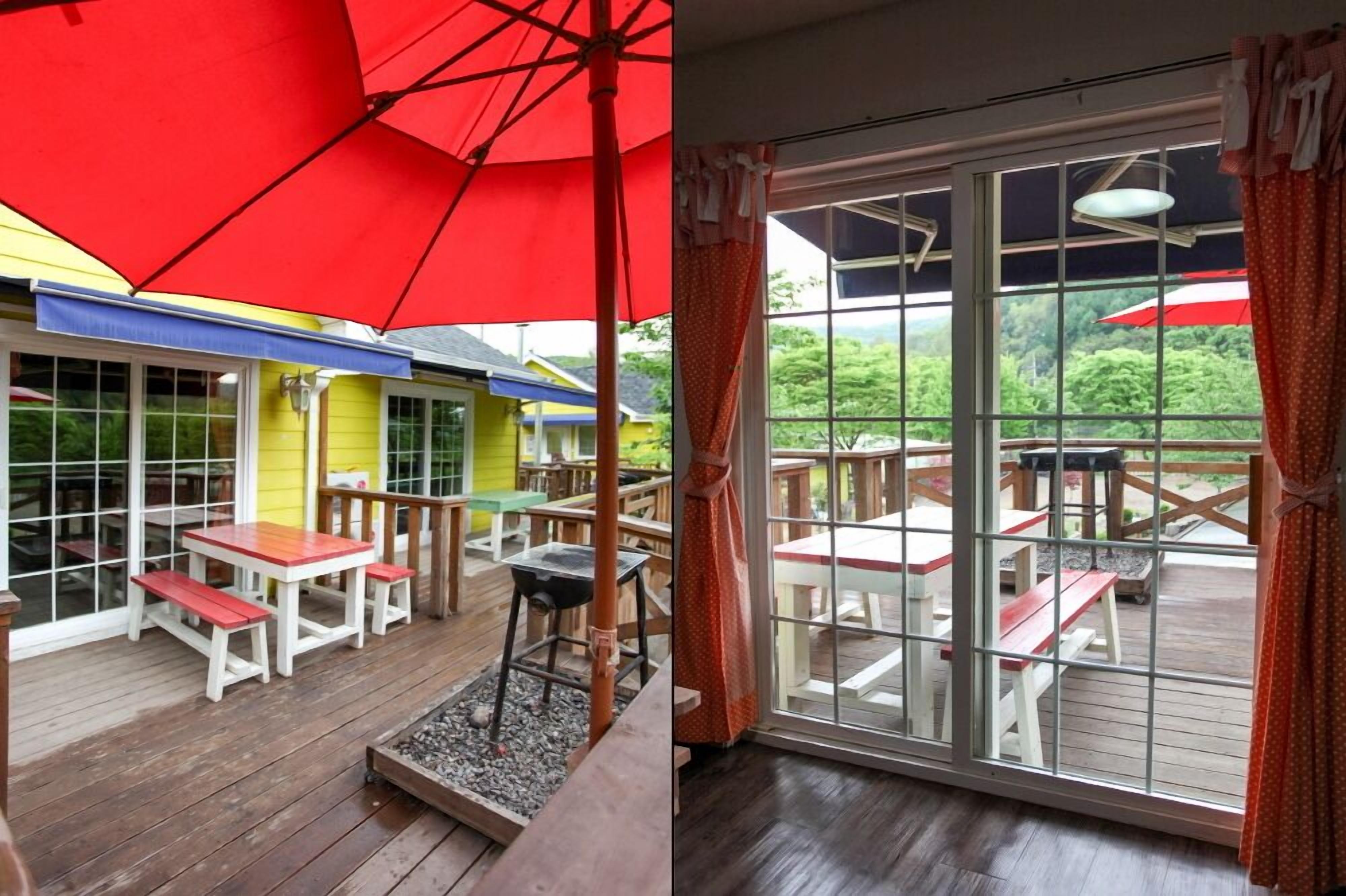 An outdoor patio with wooden tables and benches, a red umbrella, and a view of a yellow building through large windows.