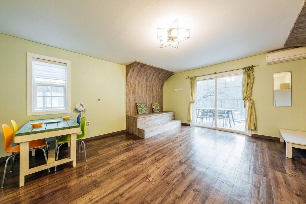 A spacious room with a wooden floor, a dining table with chairs, a bench, a staircase, and a large window with curtains.
