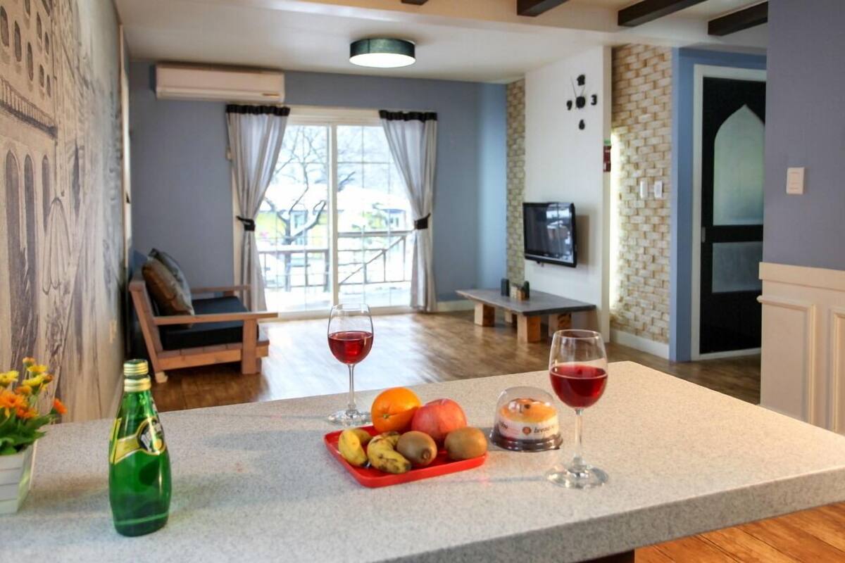 A kitchen counter with fruit and wine glasses, a bottle of soda, and a view of a living room with a sofa and a TV.