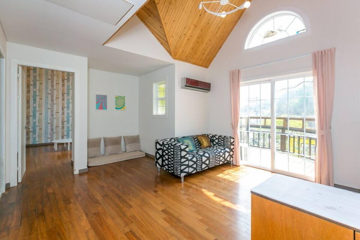 A spacious living room with a wooden ceiling, a black and white patterned sofa, a white bench, and a large window with a view of a balcony.