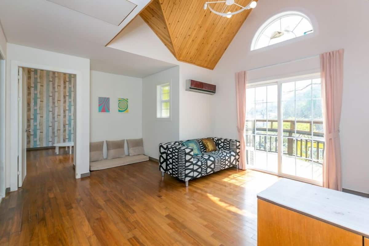 A spacious living room with a wooden ceiling, a black and white patterned sofa, a white bench, and a large window with a view of a balcony.