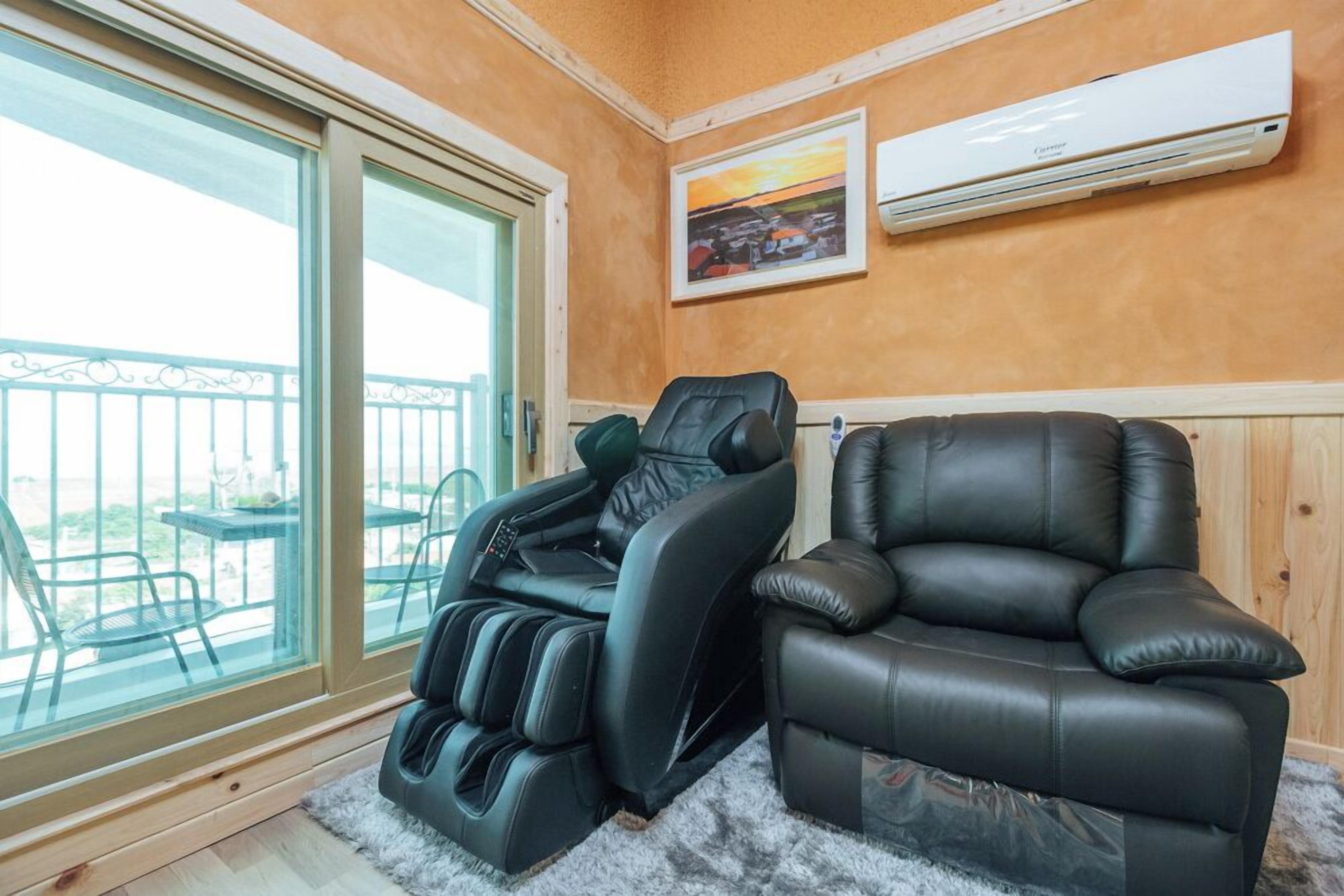 Una silla de masaje y un sillón en una habitación con un ventanal grande, un cuadro en la pared y un equipo de aire acondicionado.