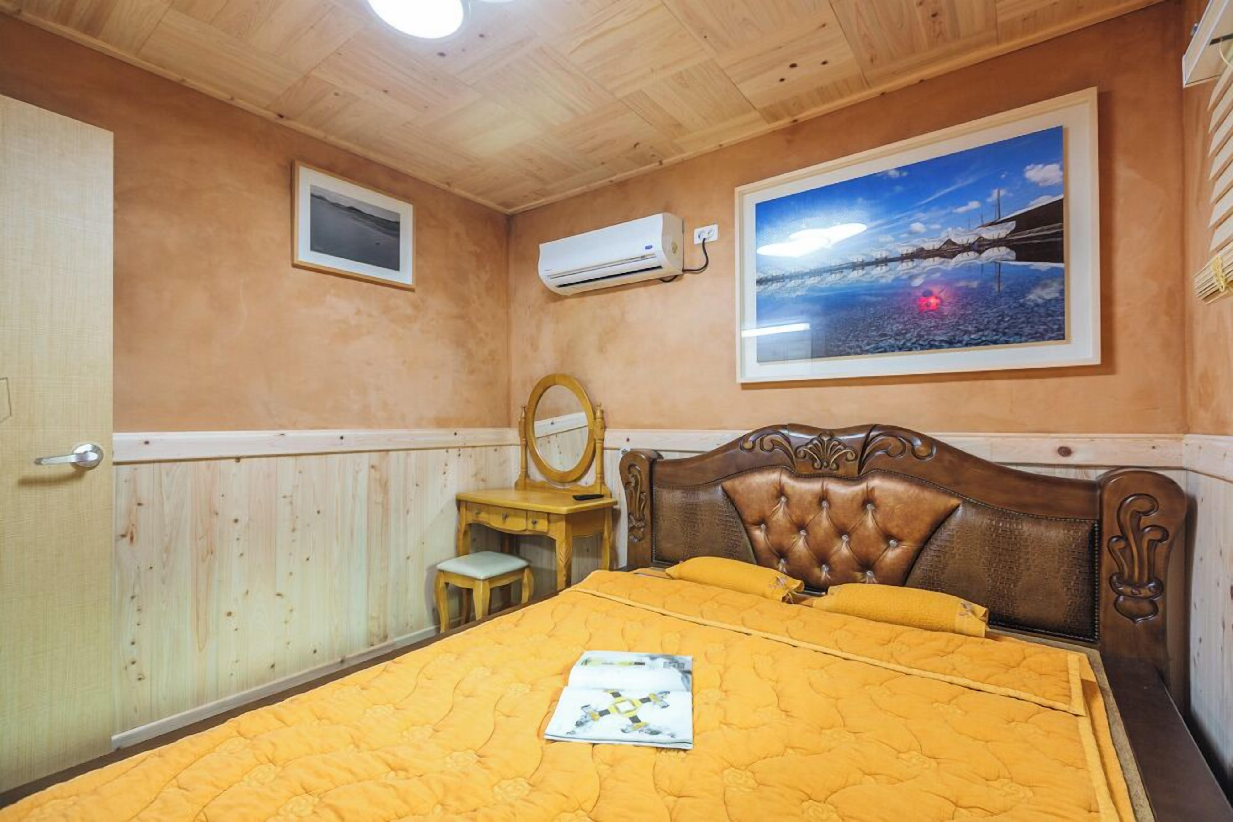 A bedroom with a wooden ceiling, a bed with a yellow quilt, a wooden headboard, a bedside table with a mirror, and a wall-mounted air conditioner.
