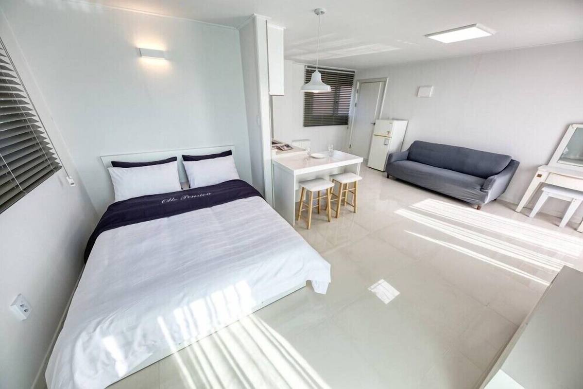 A modern hotel room with a bed, a desk, a sofa, and a small kitchenette.