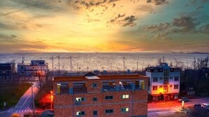 A multi-story building with a flat roof, located near a waterfront with a sunset view. - Jebudo Olle Pension (Hwaseong)