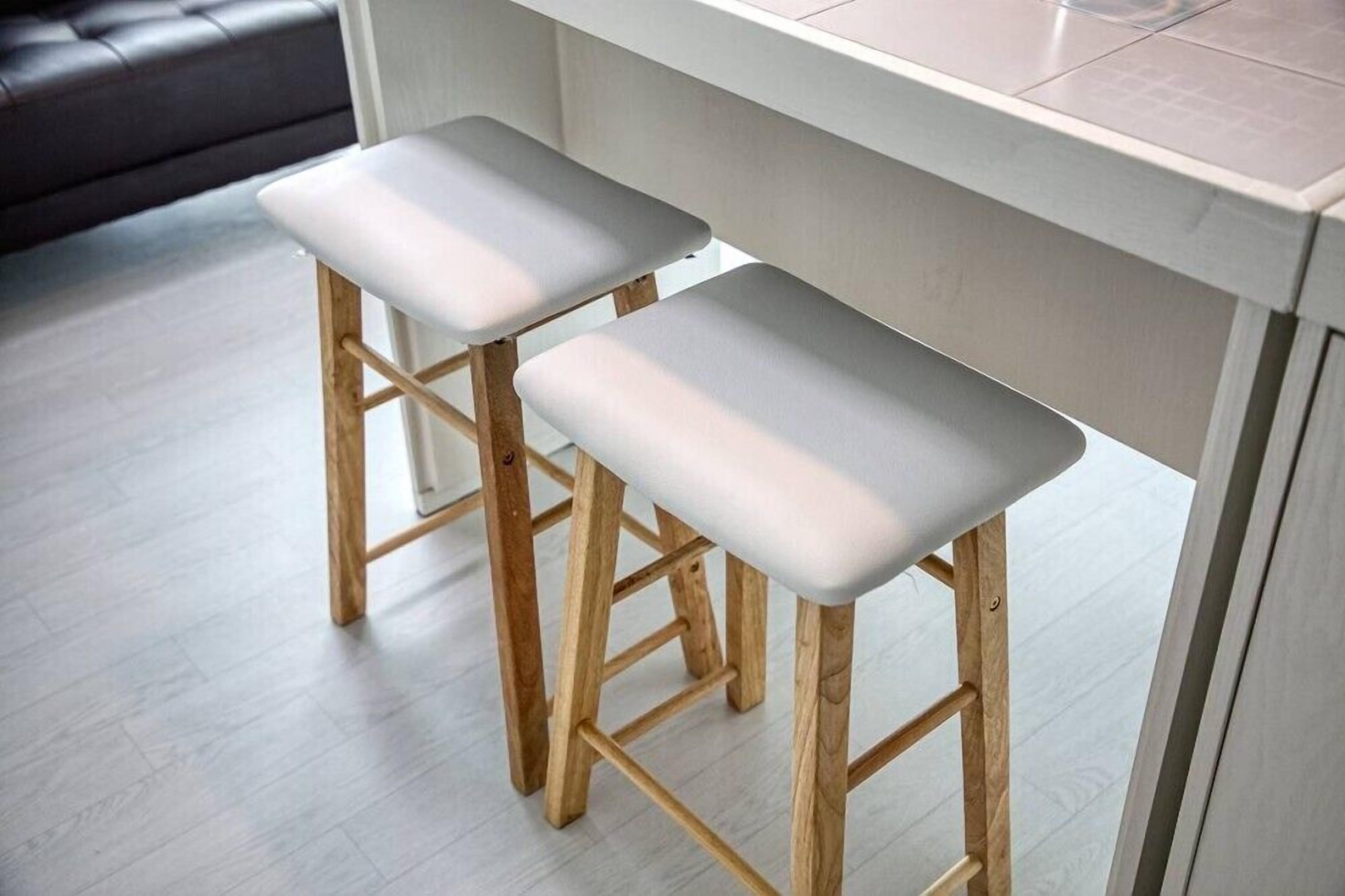 Dos taburetes de madera con cojines blancos, una encimera de baldosas de color claro y un sofá gris al fondo.