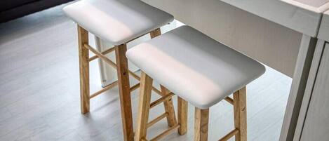 Dos taburetes de madera con cojines blancos, una encimera de baldosas de color claro y un sofá gris al fondo.