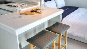 A compact kitchen area with a white countertop, two stools, a wooden cutting board with pastries, and a wine glass. - Jebudo Olle Pension (Hwaseong)